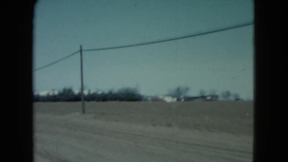 1960 Nevada rural roadside view with flat farmland, a utility pole, overhead line, and small houses on the horizon.