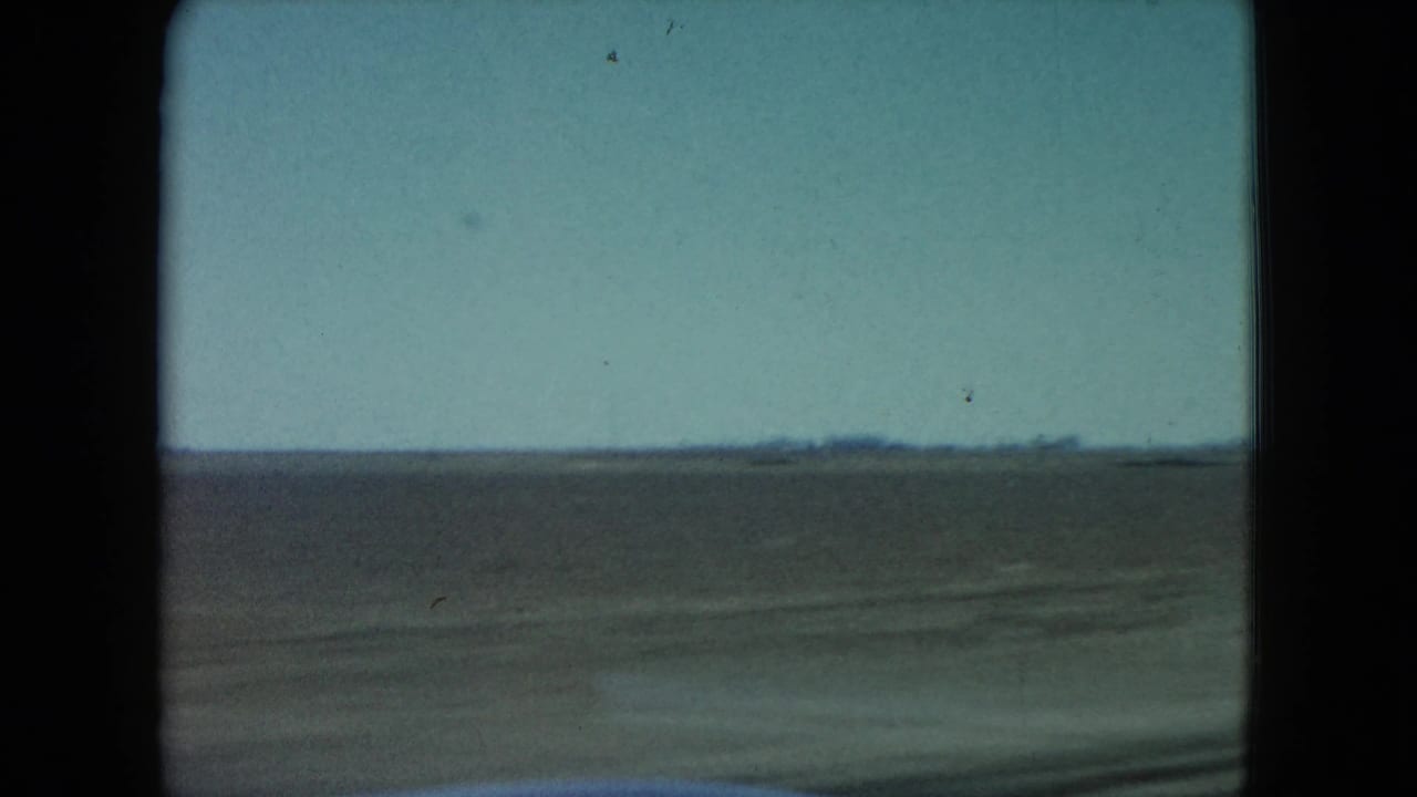 1960 Nevada home-movie frame showing arid open land through a moving car window, with a low horizon and pale blue sky.