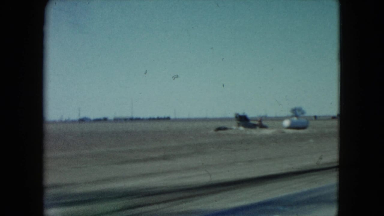 Nevada moving-car frame showing open land, distant trees, and a small aircraft-like object beside the road under a pale sky.