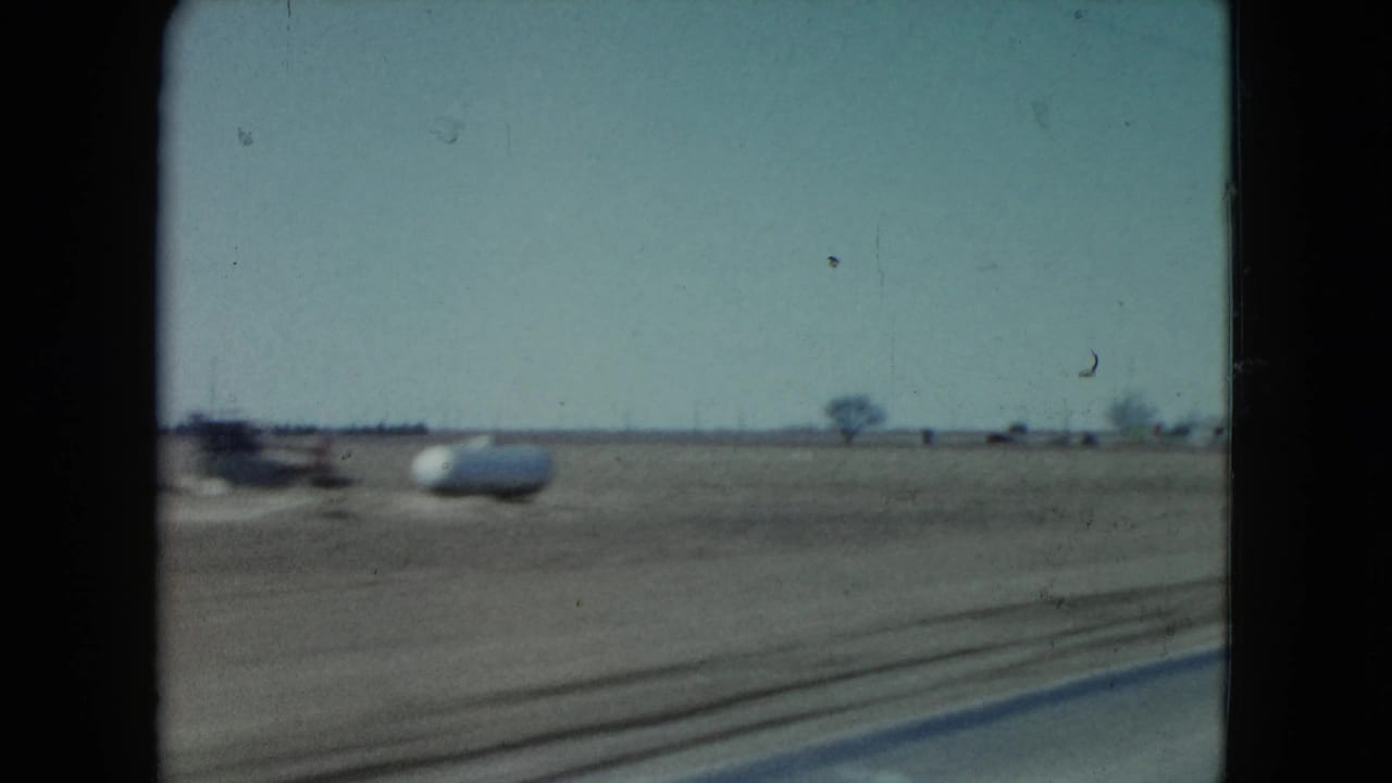 1960 Nevada home-movie frame showing a flat roadside plain through dark vehicle-window edges, with a distant light object near the horizon.