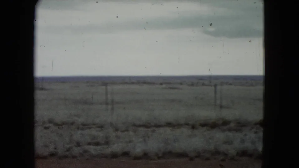Flat Nevada landscape viewed from a vehicle, with sparse fence posts, low horizon, pale sky, and visible 8mm film wear.