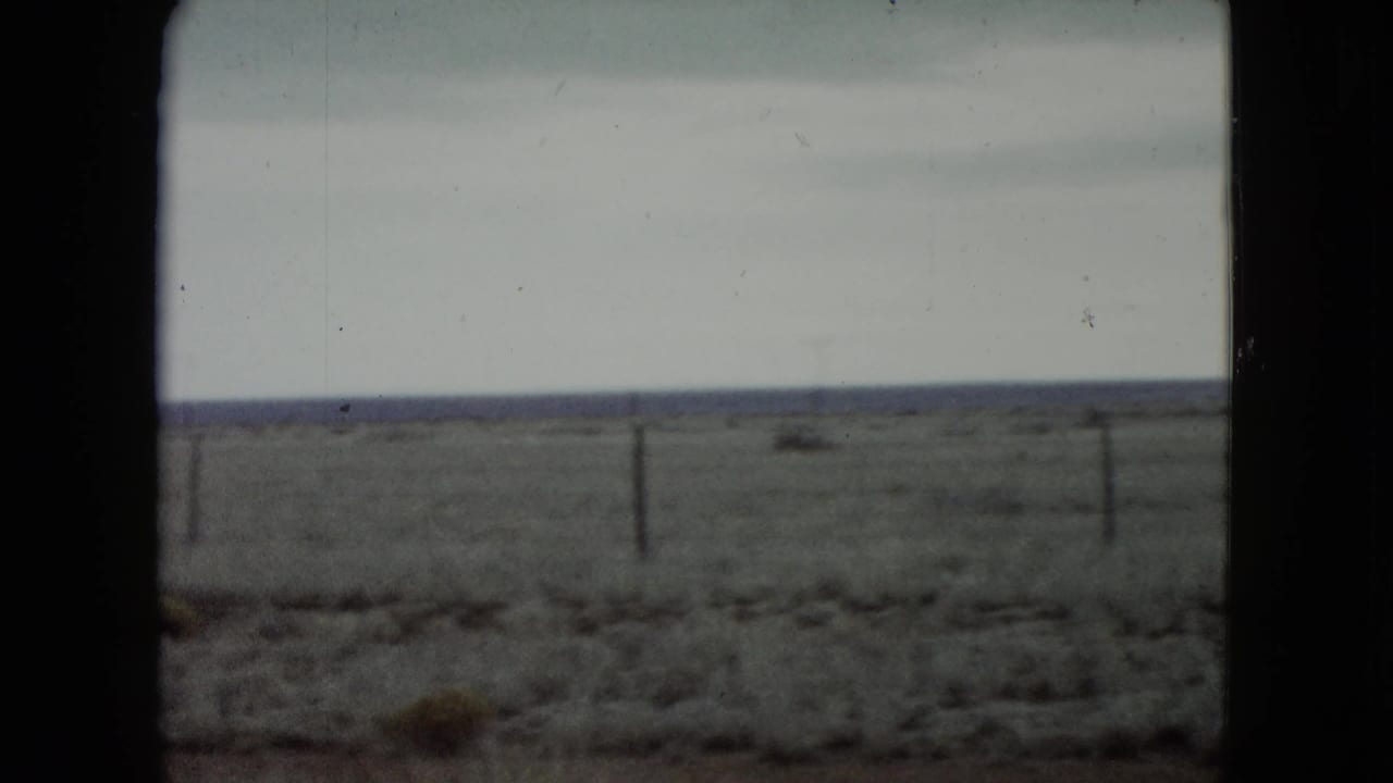 Passenger-side view across rural Nevada range, with fence posts, scrubby ground, a low distant horizon, and soft motion blur from travel.