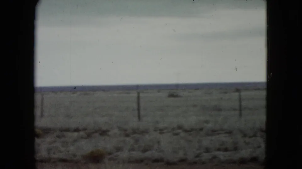 Passenger-side view across rural Nevada range, with fence posts, scrubby ground, a low distant horizon, and soft motion blur from travel.