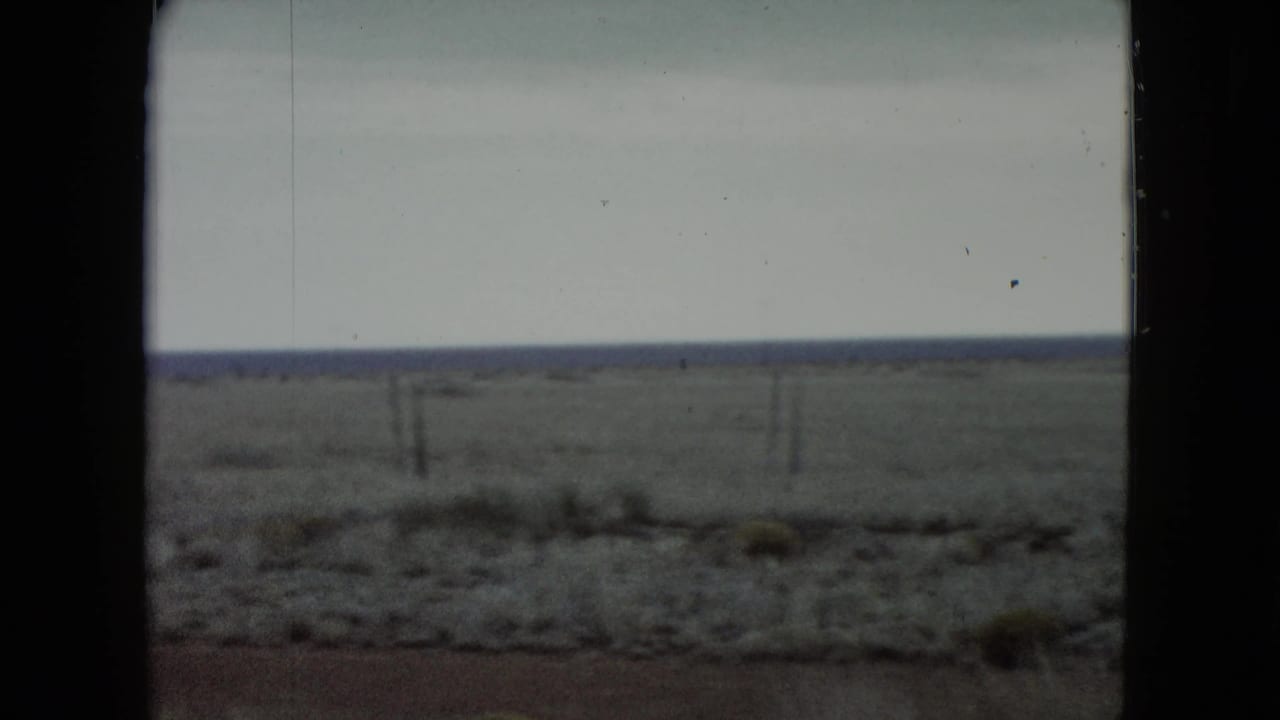 Final 1960 Nevada road-view frame showing a fenced plain, scrubby roadside foreground, low horizon, and dark film or window borders.