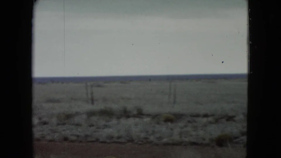 Final 1960 Nevada road-view frame showing a fenced plain, scrubby roadside foreground, low horizon, and dark film or window borders.