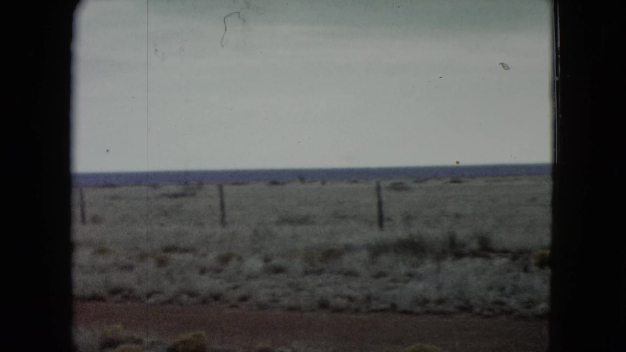 1960 Nevada 8mm frame showing sparse scrubland, reddish ground, a horizontal fence line, distant horizon, and overcast sky.