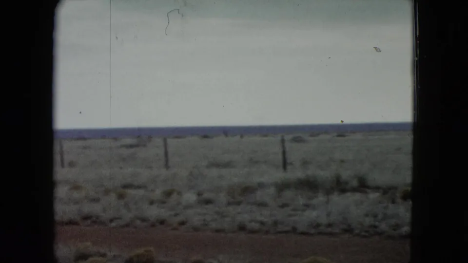 1960 Nevada 8mm frame showing sparse scrubland, reddish ground, a horizontal fence line, distant horizon, and overcast sky.