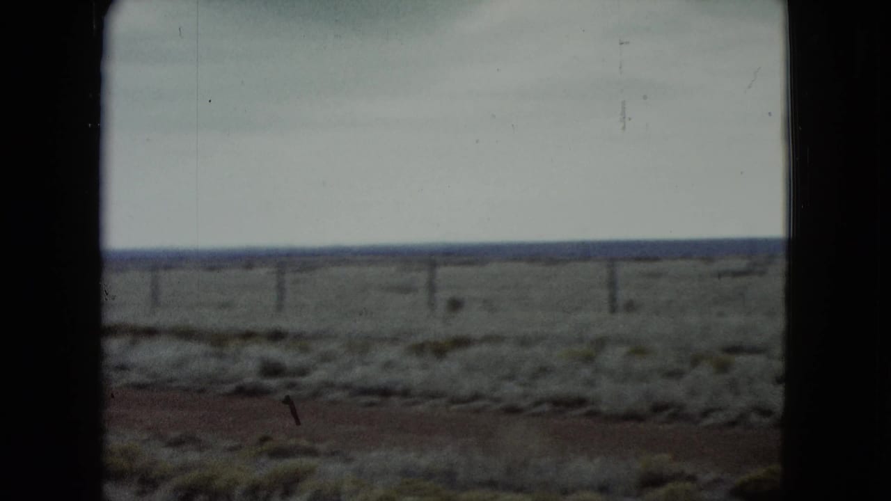 Sparse desert ground with fence posts crossing the horizon below a cloudy sky.