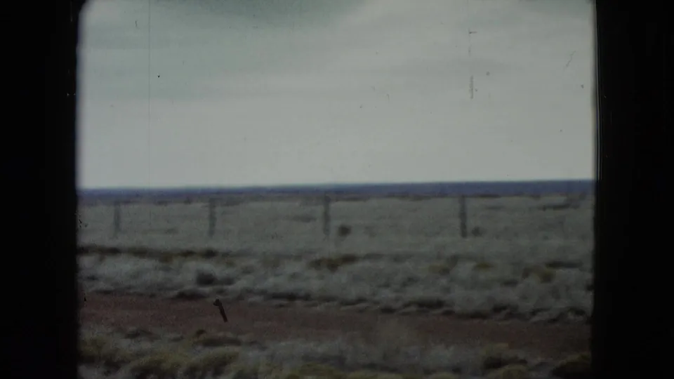 1960 Nevada travel footage frame with scrub-covered desert, a weathered fence line, open horizon, and pale cloudy sky.