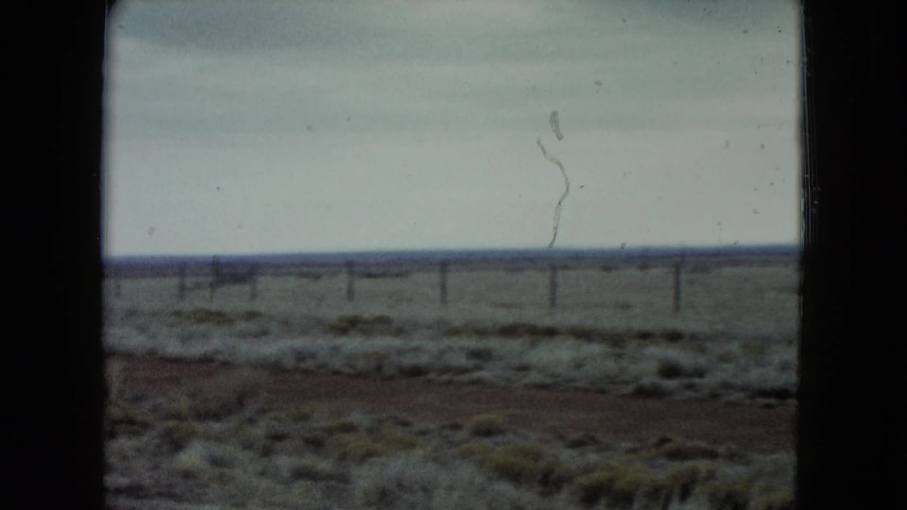1960 Nevada 8mm frame of a flat desert landscape, low vegetation, distant fence posts, and pale overcast sky.