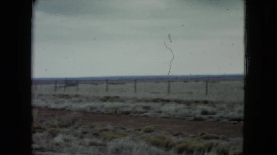 1960 Nevada 8mm frame of a flat desert landscape, low vegetation, distant fence posts, and pale overcast sky.