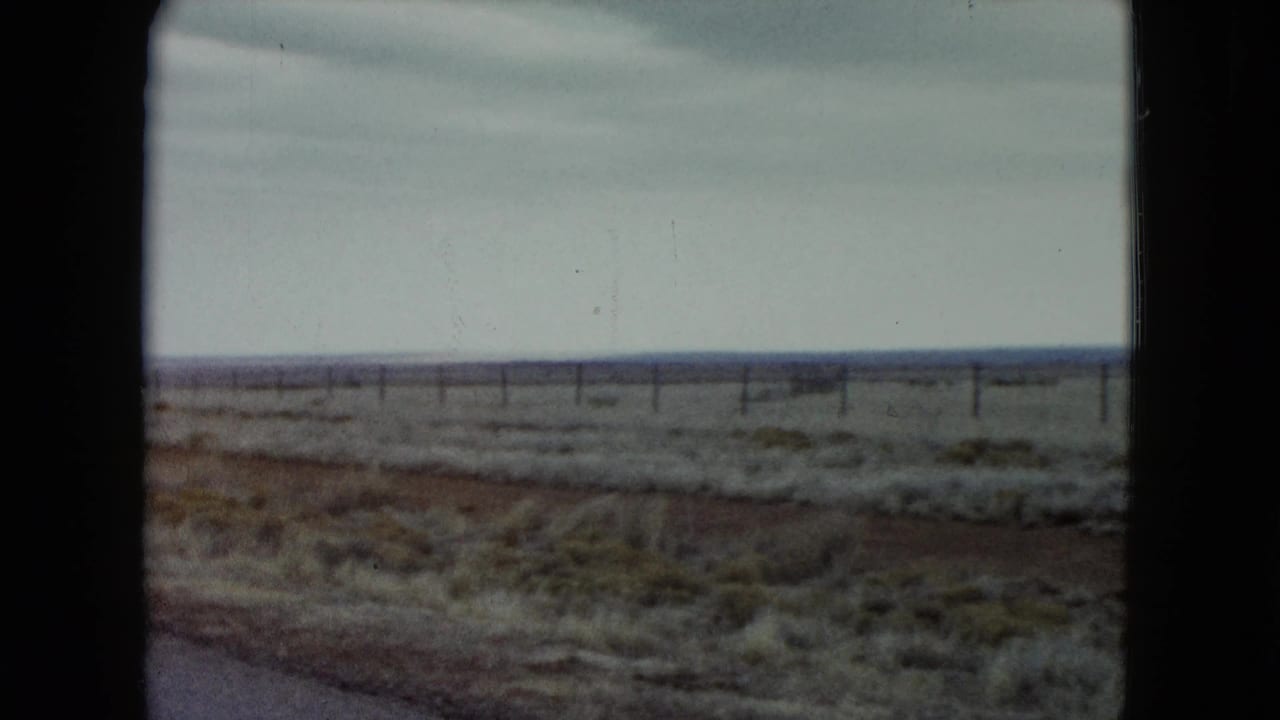 1960 Nevada roadside frame showing reddish soil, scattered scrub grass, a fence line, and a cloudy horizon from a moving vehicle.