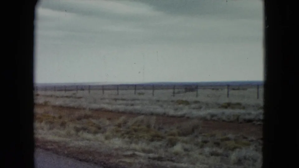 1960 Nevada roadside frame showing reddish soil, scattered scrub grass, a fence line, and a cloudy horizon from a moving vehicle.