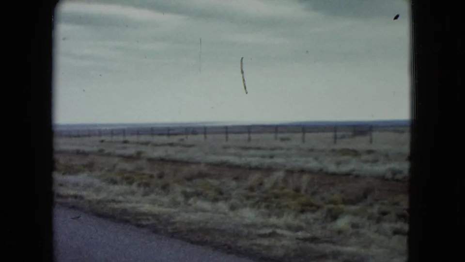 1960 Nevada home-movie view of a flat roadside plain, fence line, and cloudy sky from a moving vehicle.