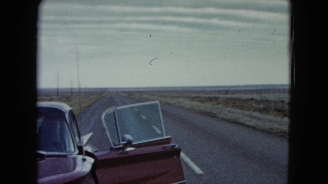 A red vintage car with an open door sits in the foreground of a 1960 Nevada road view across flat scrubland.