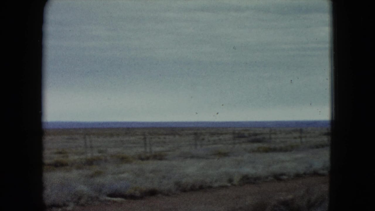 1960 Nevada 8mm frame showing a dry scrub plain, low fence posts, cloudy sky, and distant blue horizon.