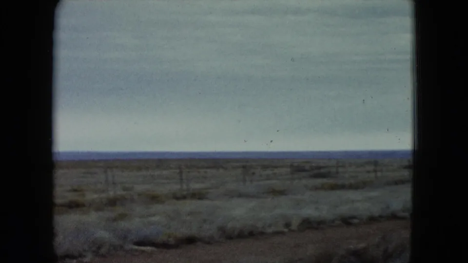 1960 Nevada 8mm frame showing a dry scrub plain, low fence posts, cloudy sky, and distant blue horizon.