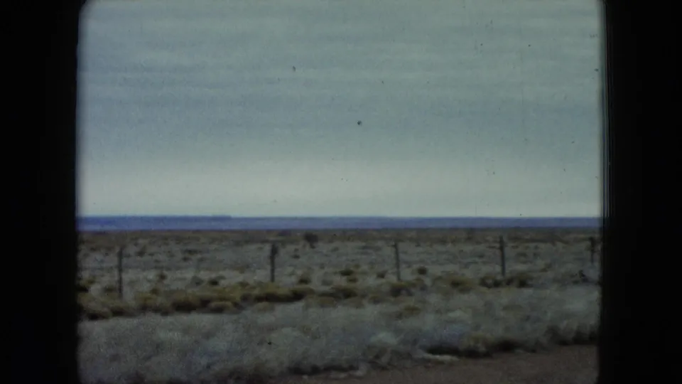 1960 Nevada home-movie frame with open scrubland, roadside fencing, and a pale cloudy sky across the horizon.