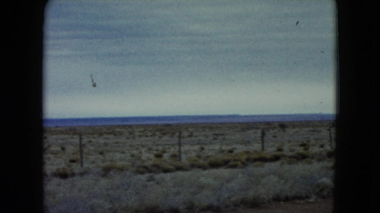 1960 Nevada 8mm view of dry grasses and scattered shrubs with fence posts set against a cloudy blue-gray horizon.