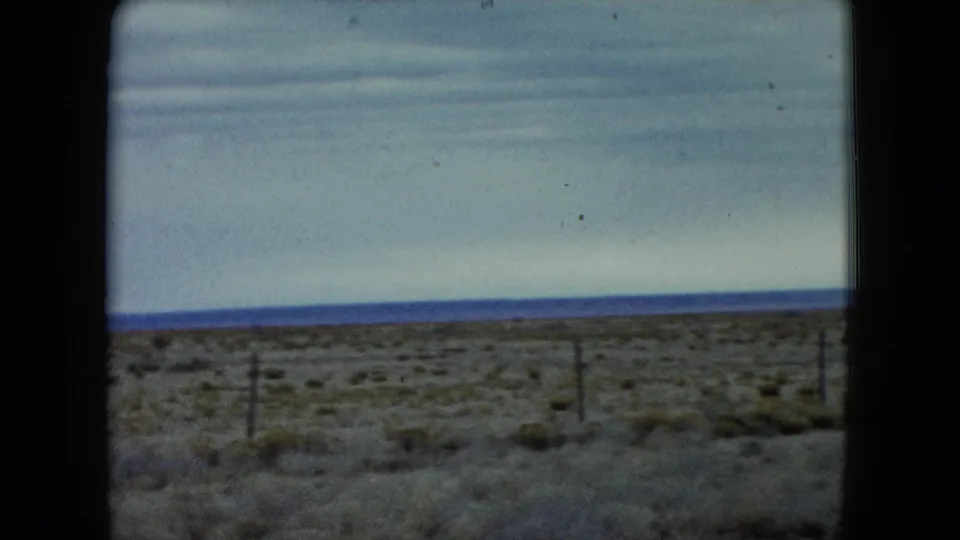 1960 Nevada 8mm frame showing fence posts, low scrub vegetation, and a broad cloudy sky over flat open land.