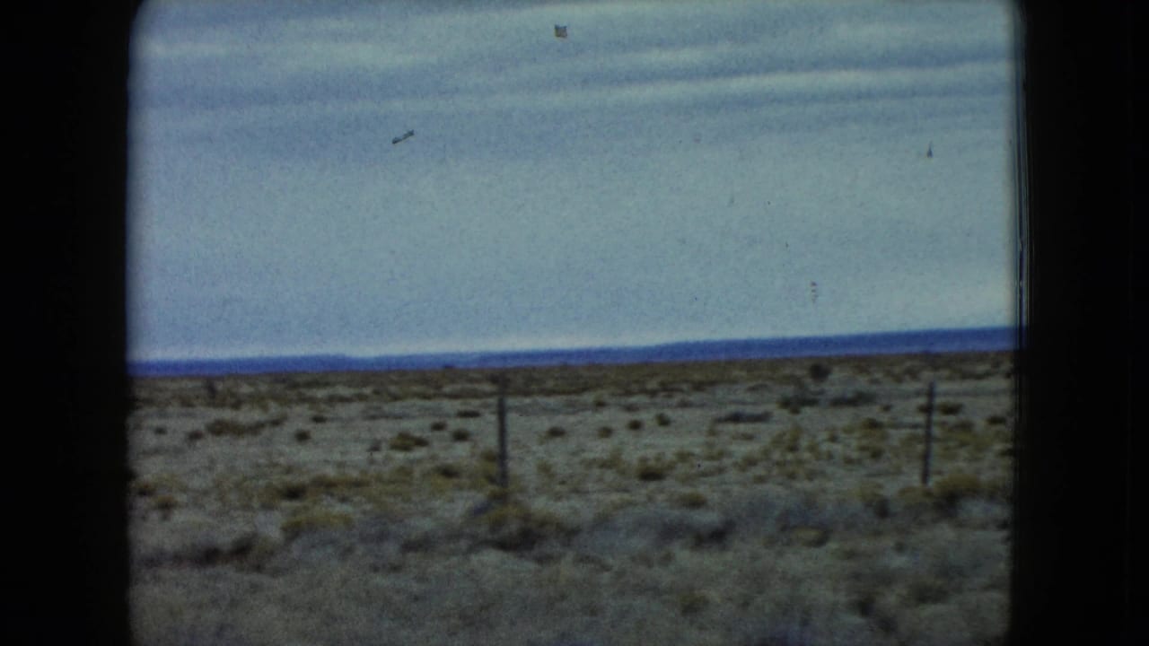 1960 Nevada home-movie frame of a broad arid landscape with scrub, fence posts, and a flat distant horizon.