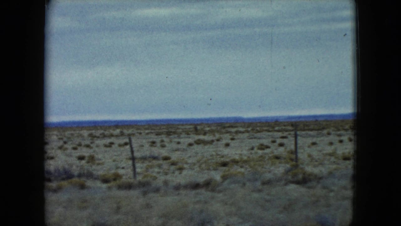 1960 Nevada 8mm frame with a tilted fence line, sparse shrubs, dry foreground, and a pale sky over the desert horizon.