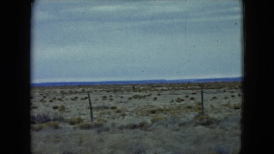 1960 Nevada 8mm frame with a tilted fence line, sparse shrubs, dry foreground, and a pale sky over the desert horizon.