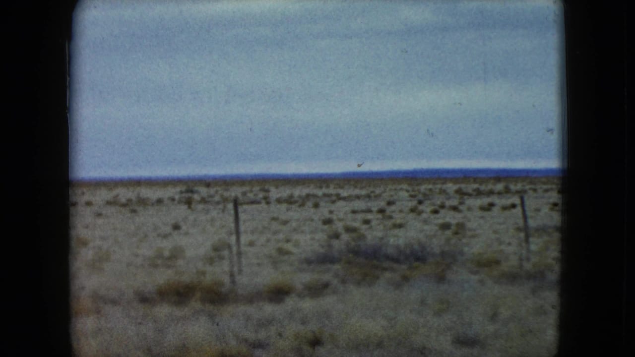 Flat desert countryside with fence posts under a muted blue sky.
