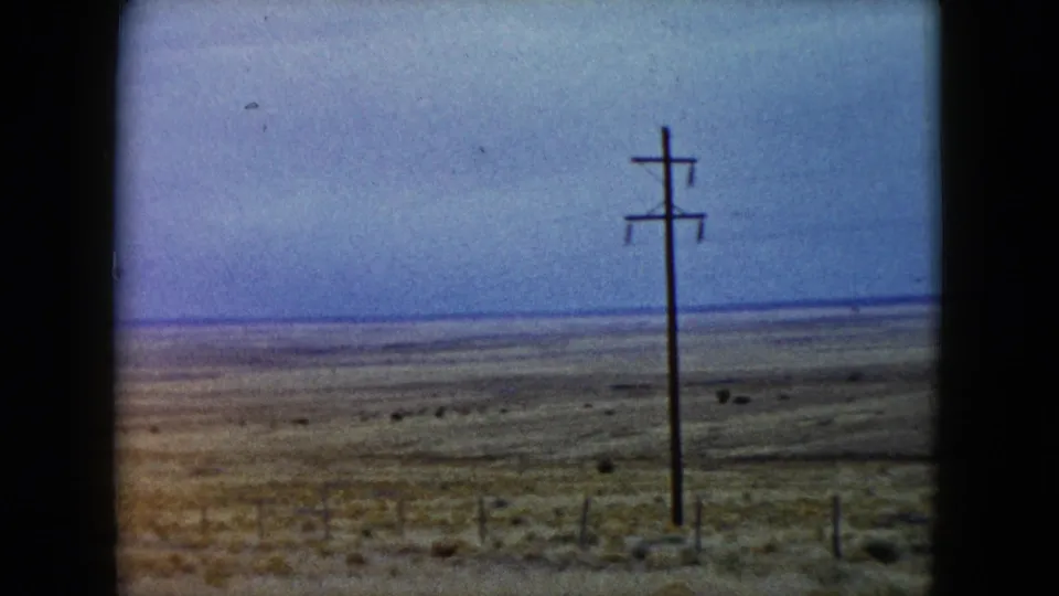 1960 Nevada roadside view with a wooden utility pole, wire fencing, sparse desert vegetation, and a broad blue-gray sky.