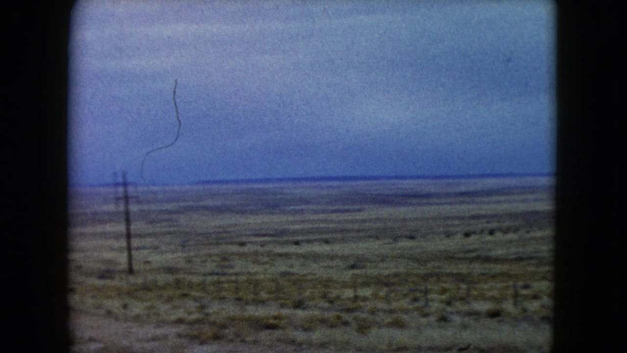 1960 Nevada 8mm frame of a wide arid plain, low brush, a roadside utility pole, and a faint curved film scratch in the sky.