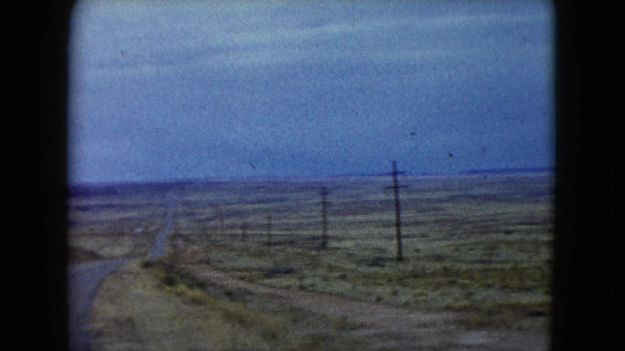 1960 Nevada driving view showing a narrow road cutting through open desert, with utility poles, fence posts, and low shrubs along the route.