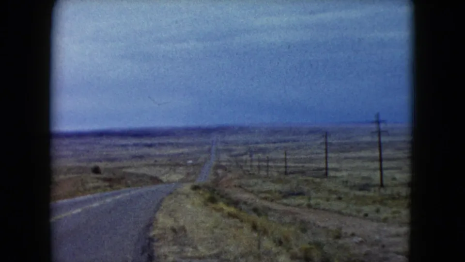 1960 Nevada 8mm frame showing an empty two-lane desert road, utility poles, sparse scrub, and a cloudy horizon.