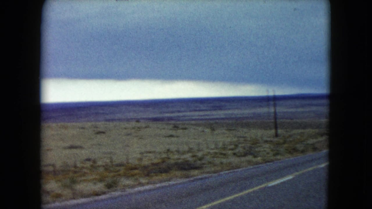 1960 Nevada 8mm frame from a moving vehicle, showing highway edge, scrubland, poles, and a bright storm-lit horizon.