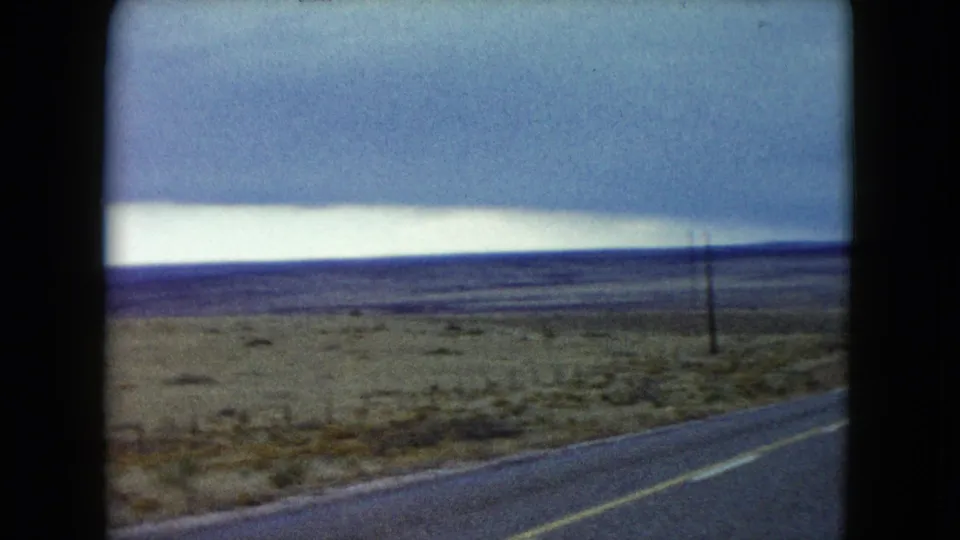 1960 Nevada 8mm frame from a moving vehicle, showing highway edge, scrubland, poles, and a bright storm-lit horizon.