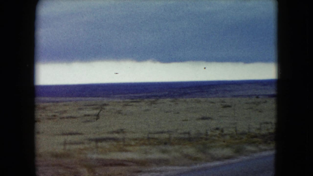 1960 Nevada 8mm frame showing flat open desert, a partial roadside view, distant terrain, and a broad cloudy sky.