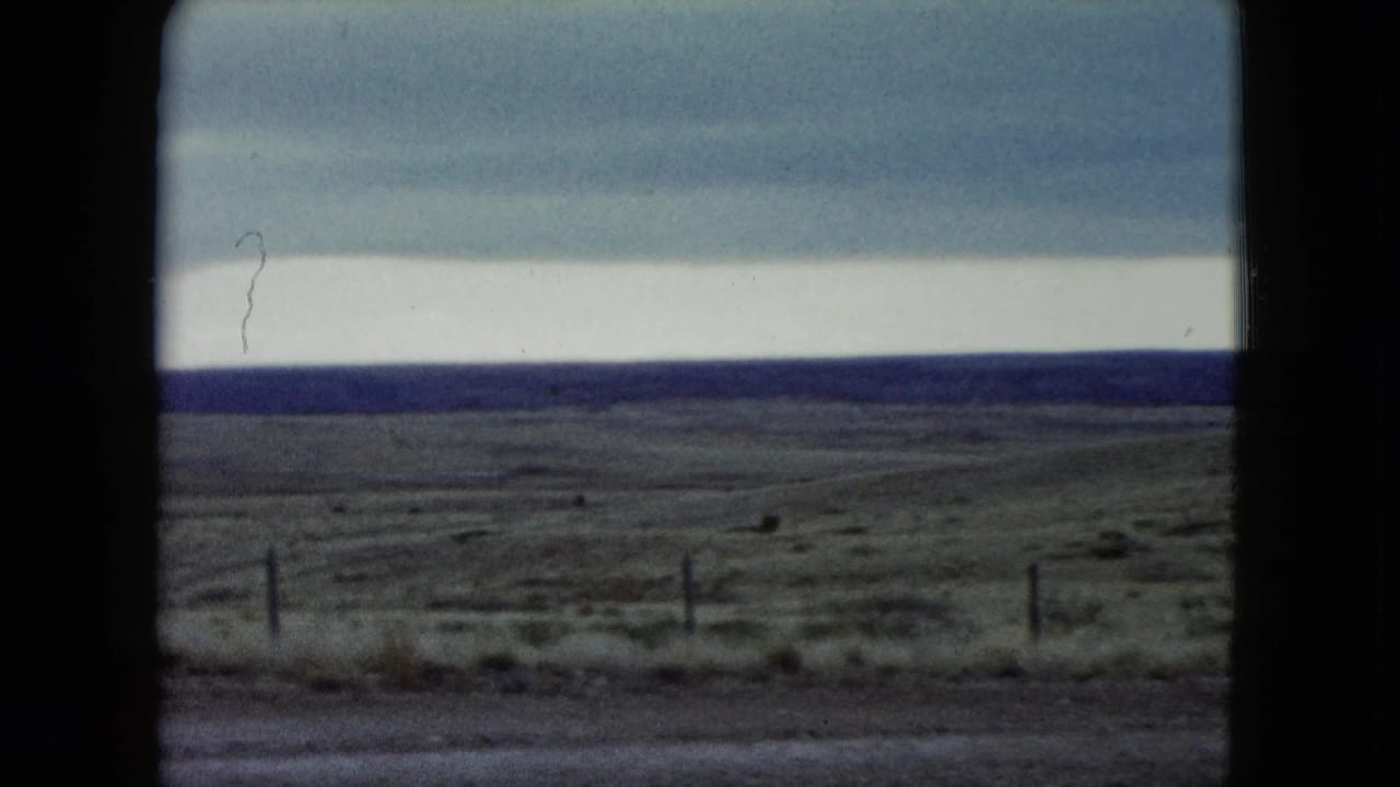 1960 Nevada home-movie frame showing a wide fenced prairie, layered overcast sky, dark horizon, and sparse roadside foreground.