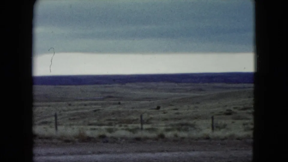 1960 Nevada home-movie frame showing a wide fenced prairie, layered overcast sky, dark horizon, and sparse roadside foreground.