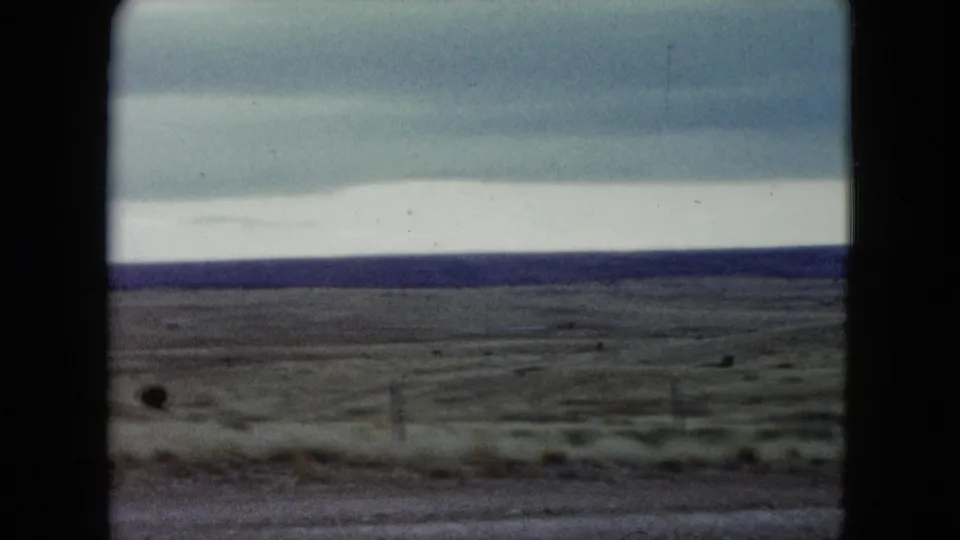 1960 Nevada 8mm frame showing dry grassland from a roadside viewpoint, with fence posts, muted sky, and a flat horizon.
