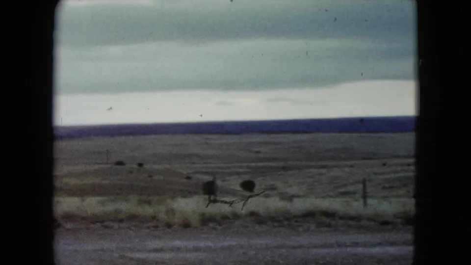 1960 Nevada home-movie frame of sparse roadside grassland, low fencing, and a wide cloudy sky over a distant horizon.
