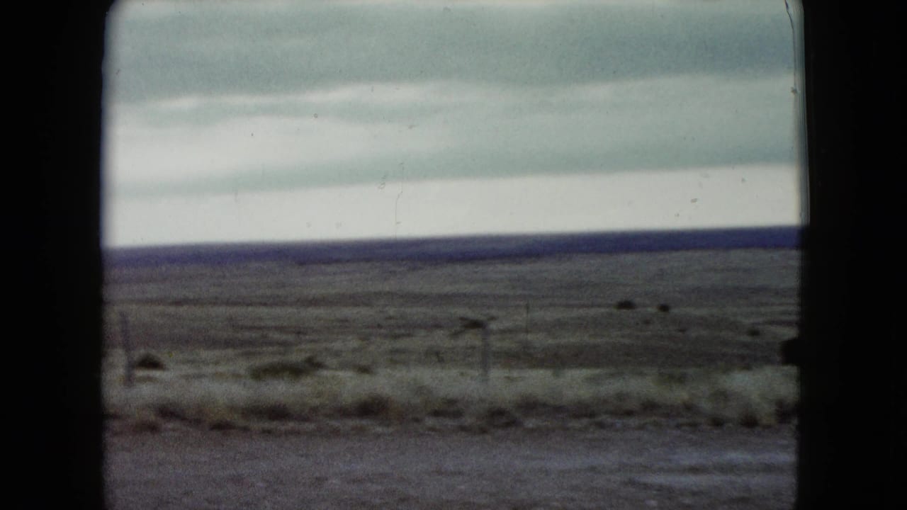 Moving 8mm view of Nevada grassland in 1960, with a blurred roadside edge, low fence posts, and layered clouds.