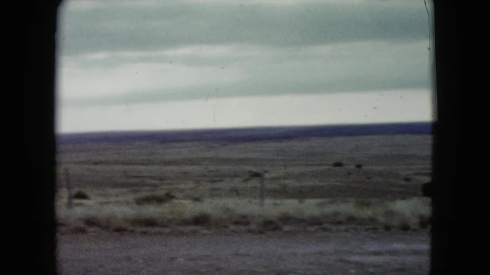 Moving 8mm view of Nevada grassland in 1960, with a blurred roadside edge, low fence posts, and layered clouds.