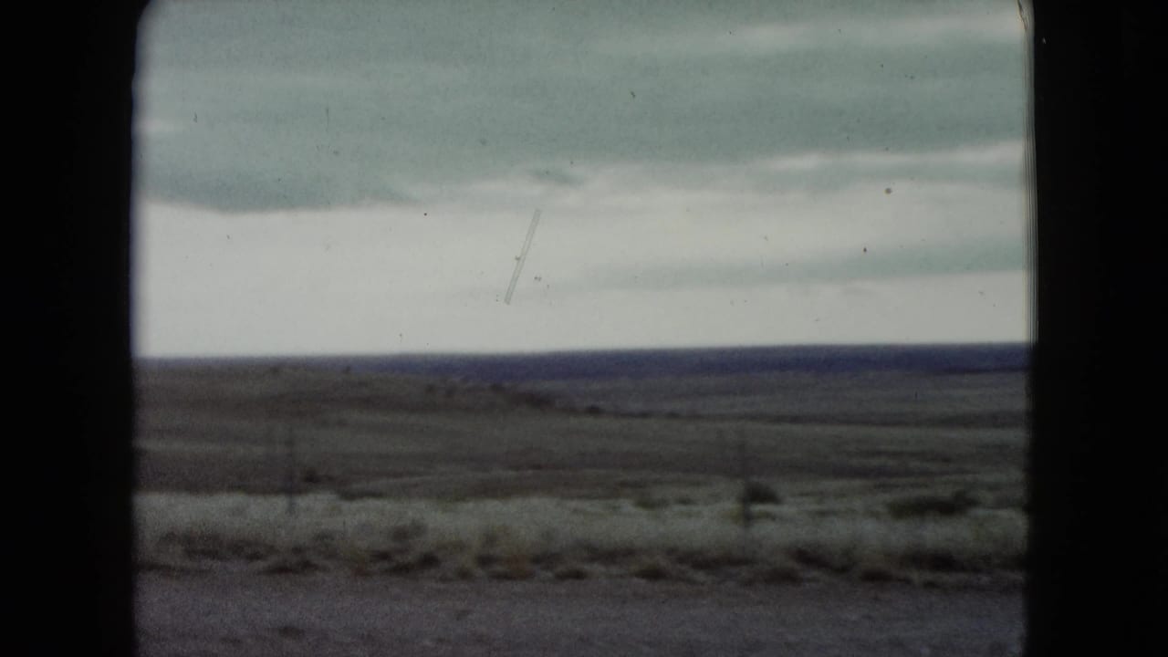 1960 Nevada 8mm frame with a broad dry plain, pale cloudy sky, dark distant horizon, and soft roadside grasses.