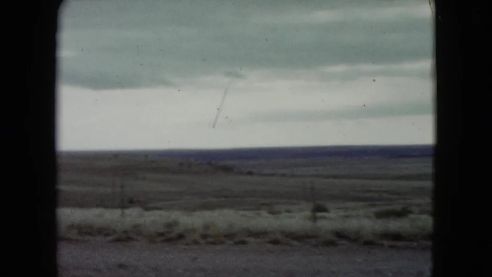1960 Nevada 8mm frame with a broad dry plain, pale cloudy sky, dark distant horizon, and soft roadside grasses.