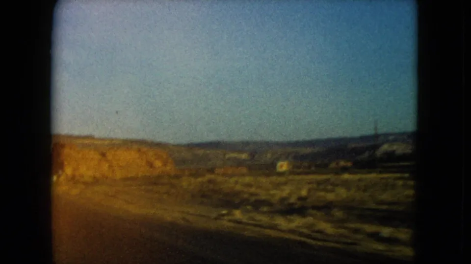 Moving 8mm view of a Nevada desert roadside with rocky formations, open sky, and a small distant structure-like shape.