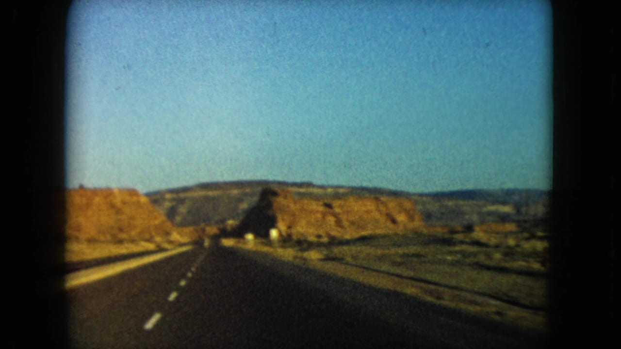 1960 home-movie view down an empty Nevada highway toward low desert plateaus and rock cuts.