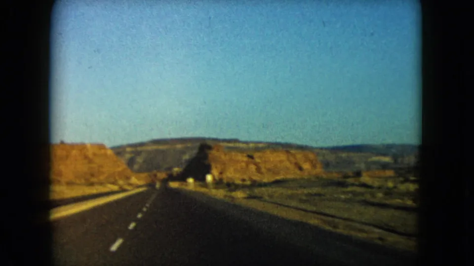 1960 home-movie view down an empty Nevada highway toward low desert plateaus and rock cuts.