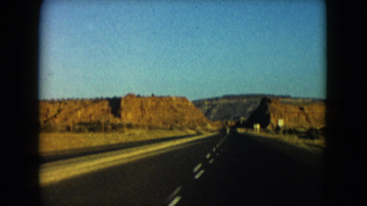 Forward-facing 8mm frame of a Nevada highway passing between rugged desert rock cuts under a clear sky.