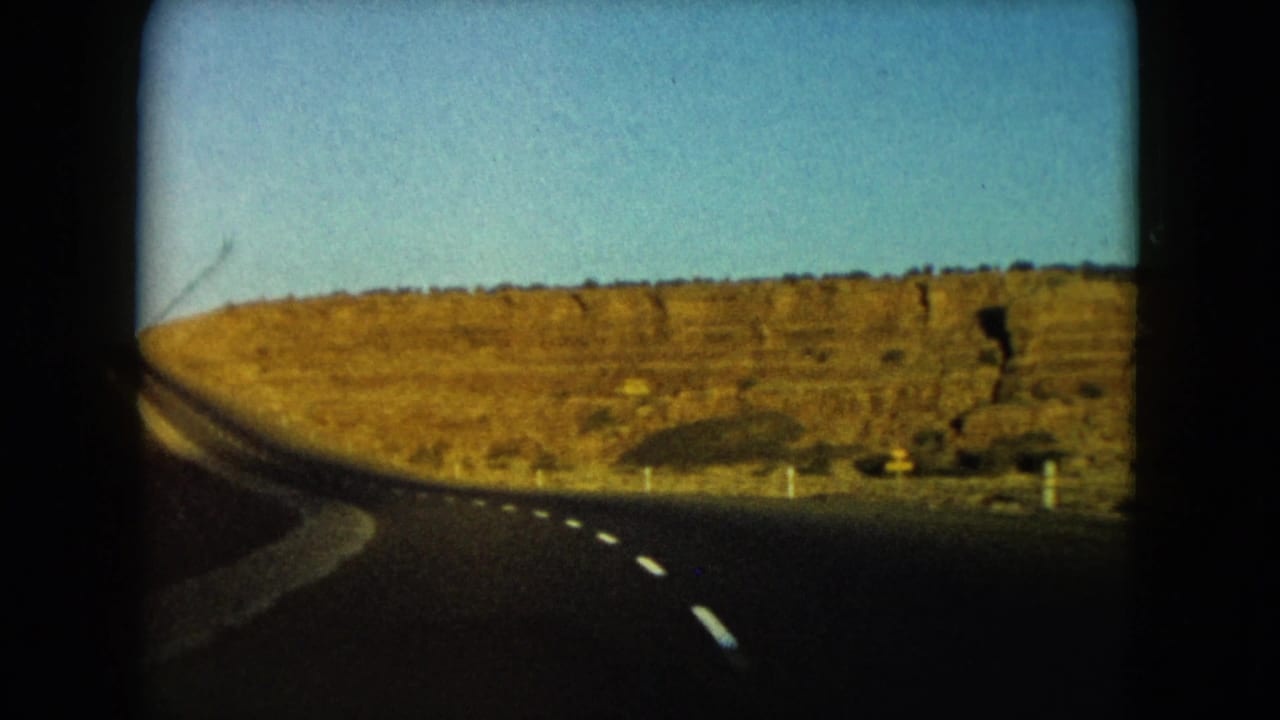 1960 Nevada road-trip footage frames a bend in the highway below a broad rocky desert hillside.