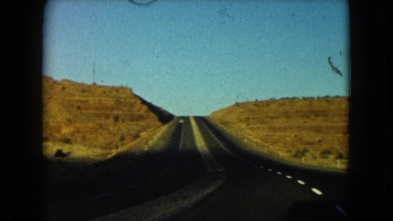 1960 Nevada 8mm frame with a straight highway climbing between dry rocky hills under clear blue sky.