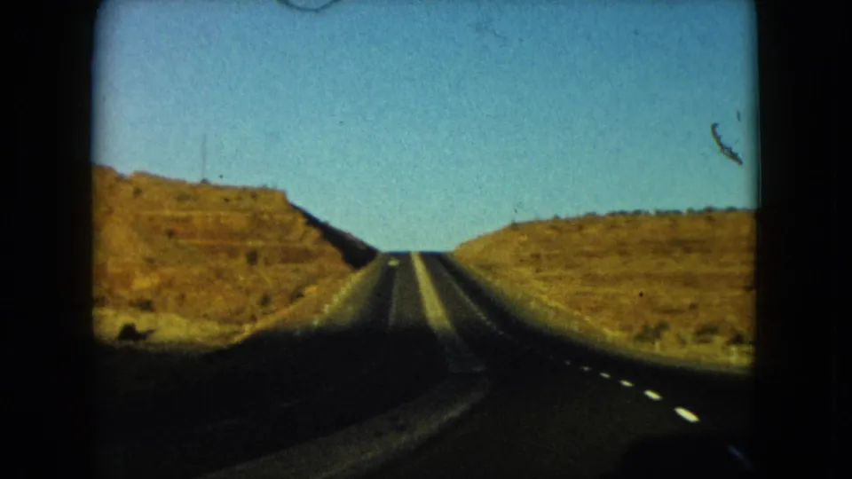 1960 Nevada 8mm frame with a straight highway climbing between dry rocky hills under clear blue sky.